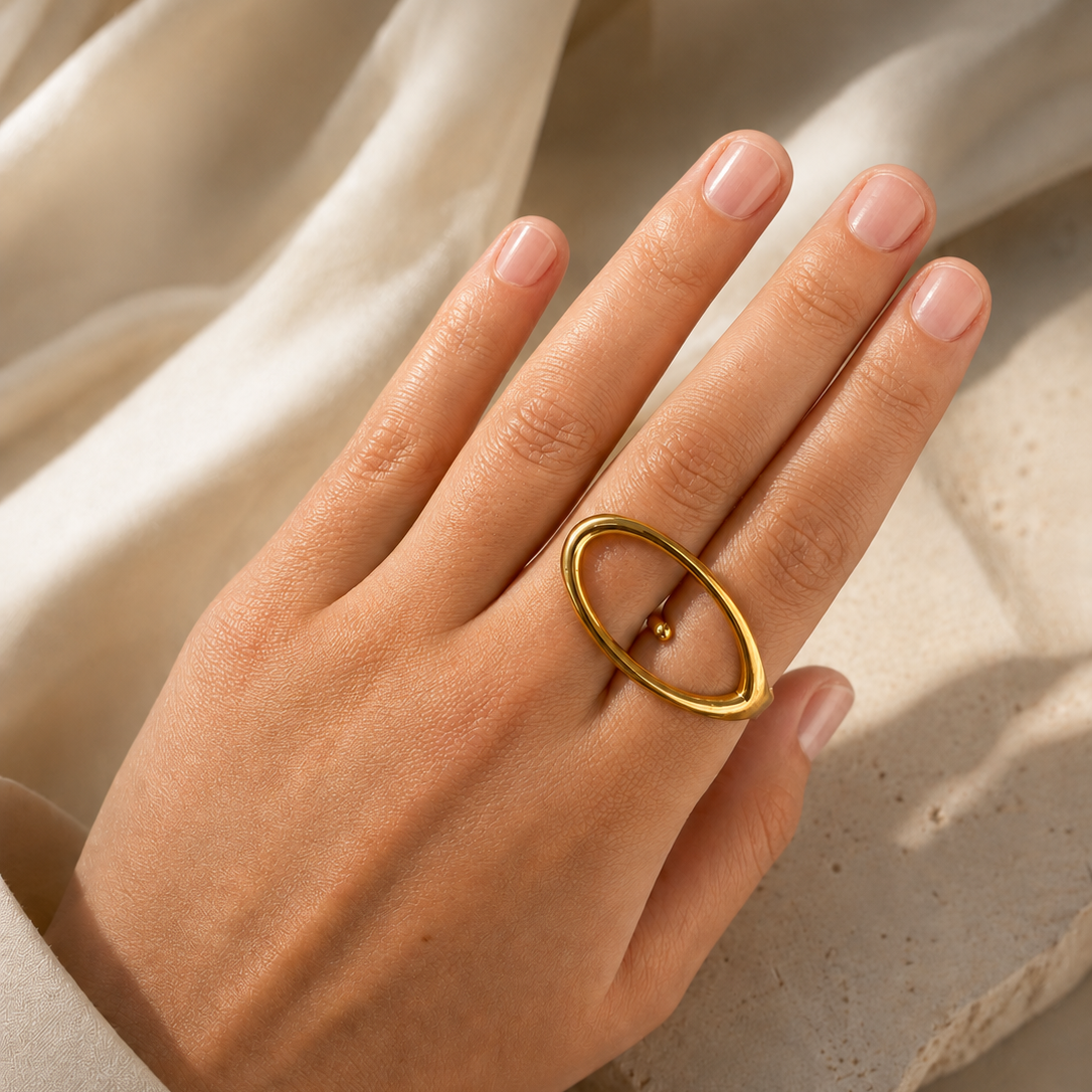 Model wears the gold Aura RING's large open oval loop on her finger against a draped ivory fabric.