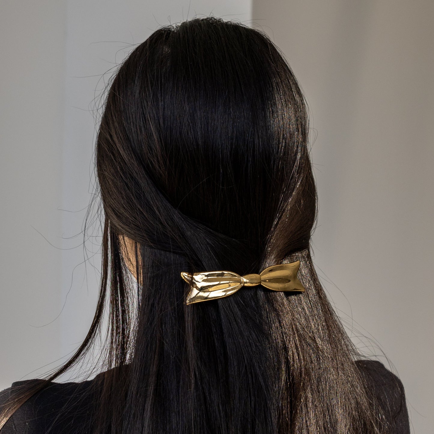 Bow HAIR CLIP in gold-tone metal, worn low on straight dark hair, close-up back view.
