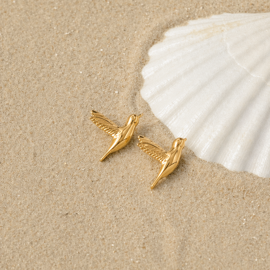 Two Kai EARRINGS hummingbird studs rest on sand beside a white seashell for a soft, beach-inspired styled shot.