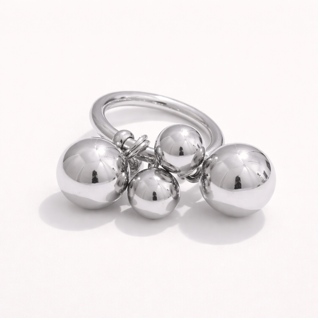 Silver ring with a cluster of four polished ball charms dangling from the band, shown at an angle on a cream backdrop.