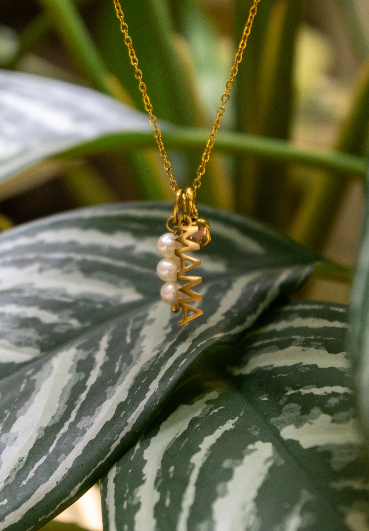 The Mama CHARM worn with a small gem and pearl accent, the gold MAMA pendant resting on a striped green houseplant leaf.