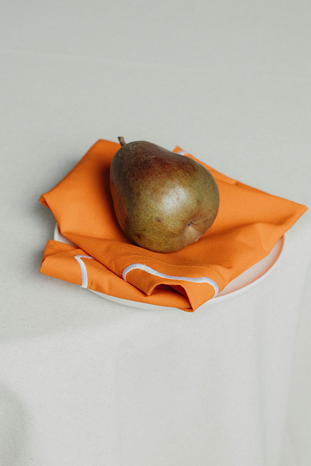 A pear resting on a folded orange Maude napkin with white wave-trim edging, set on a white plate.