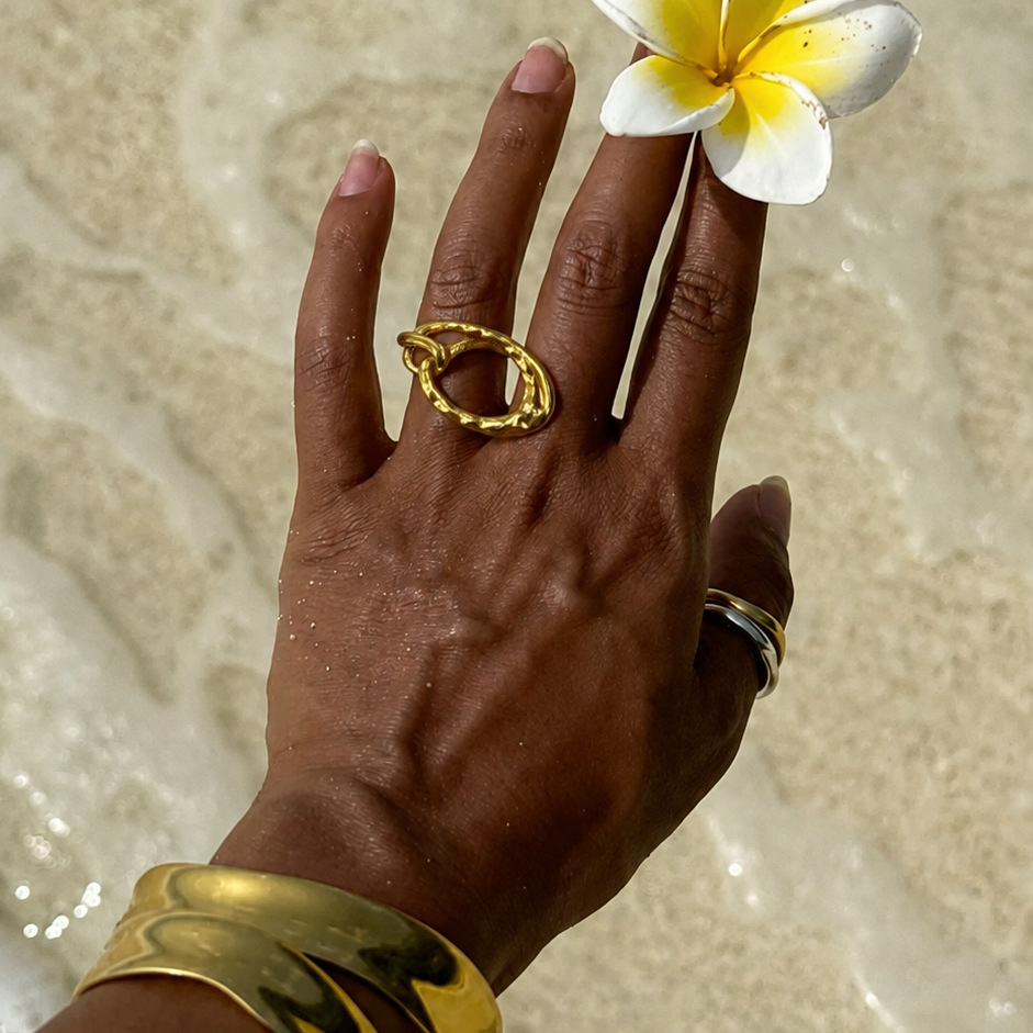 Model wears the gold Nova RING on a hand at the water's edge, a plumeria flower and stacked bangles nearby.