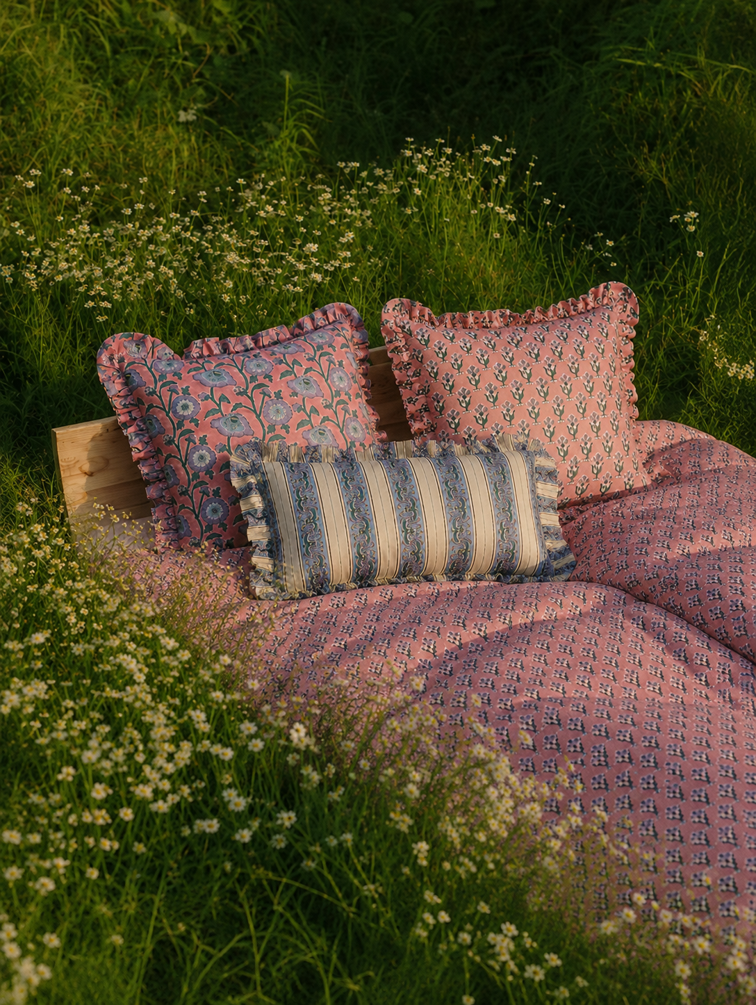 Pink patterned bedding styled with the ruffled Razia, Yavi and Taj cushions on a rustic bed set among grass and wildflowers at sunset.