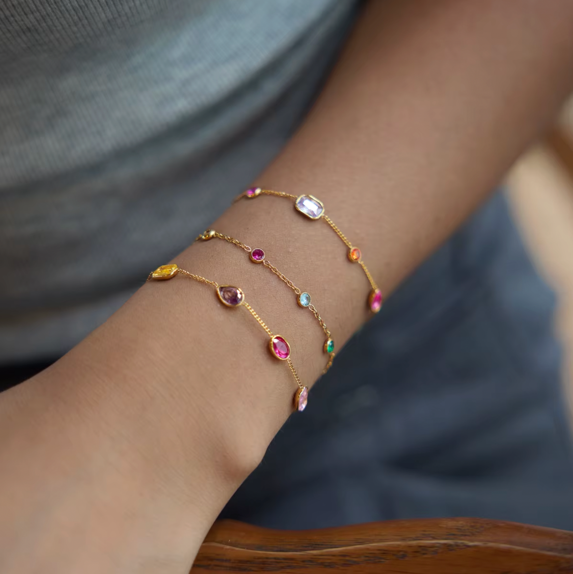 Arm resting on a wooden chair wearing three stacked gold Riva BRACELETs with multicolored oval, round and emerald-cut stones.
