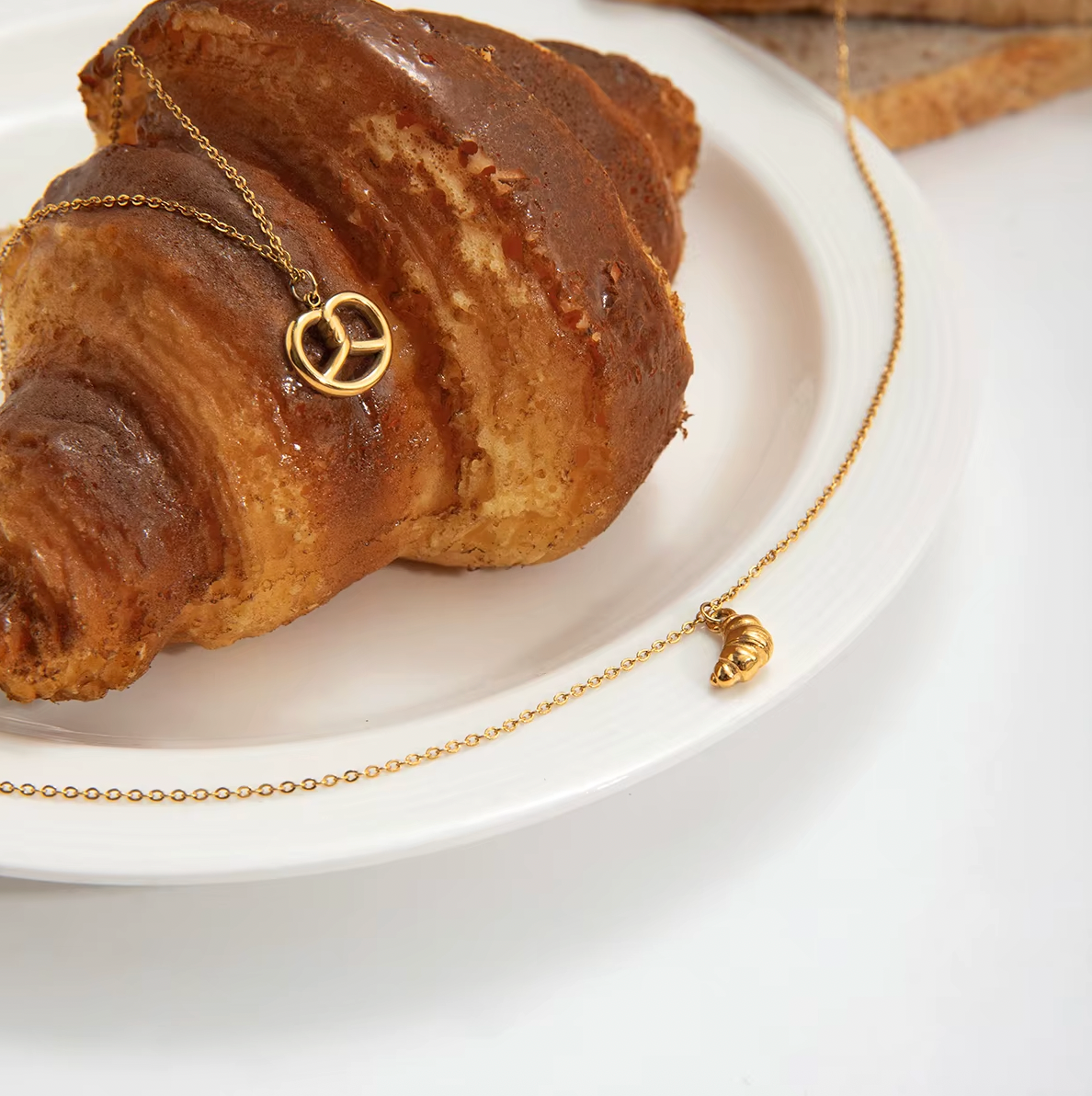 The Salty Sweet NECKLACE and its matching croissant charm necklace draped over a glazed croissant on a white plate.