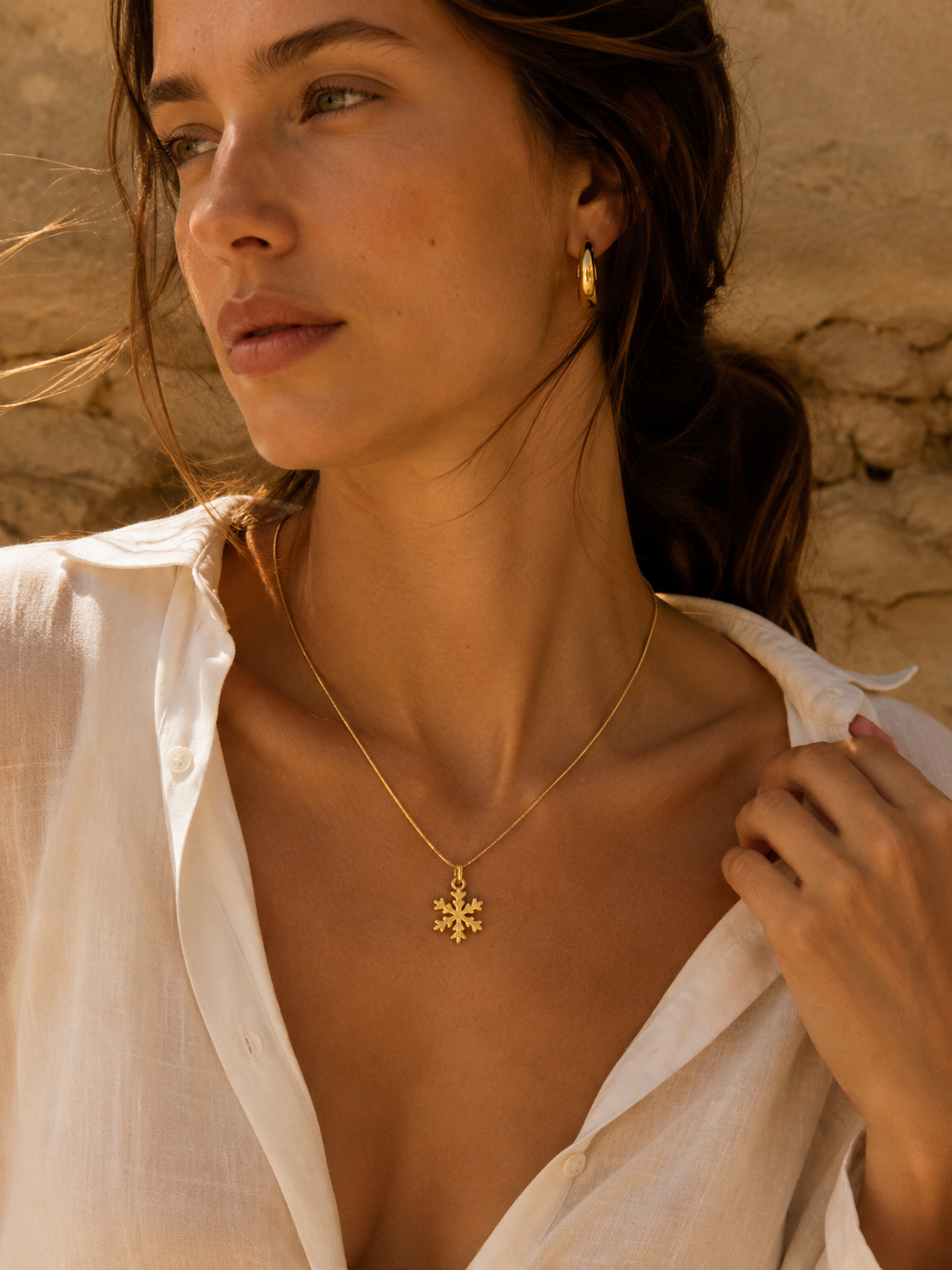 The Snowflake CHARM worn as a pendant on a fine gold chain necklace, resting at the neckline over a white linen shirt.