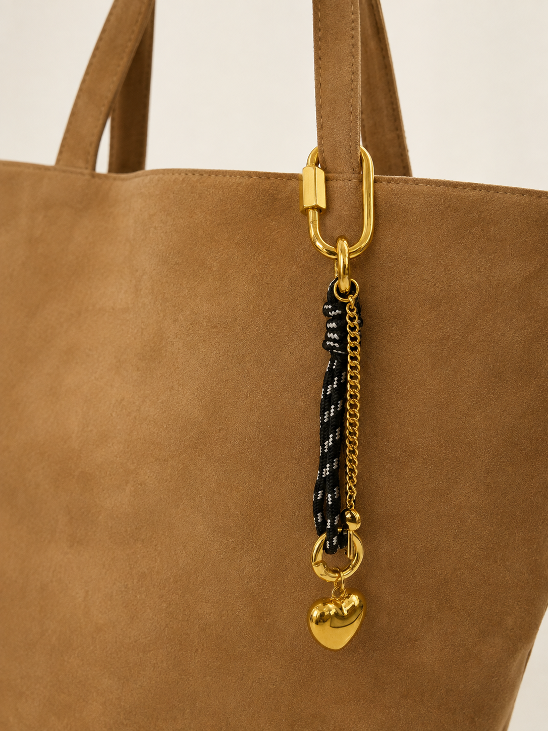 A close-up of a tan suede tote bag featuring a gold-tone lobster clasp and a hanging charm with a black-and-white braided rope, a gold chain, and a gold heart.