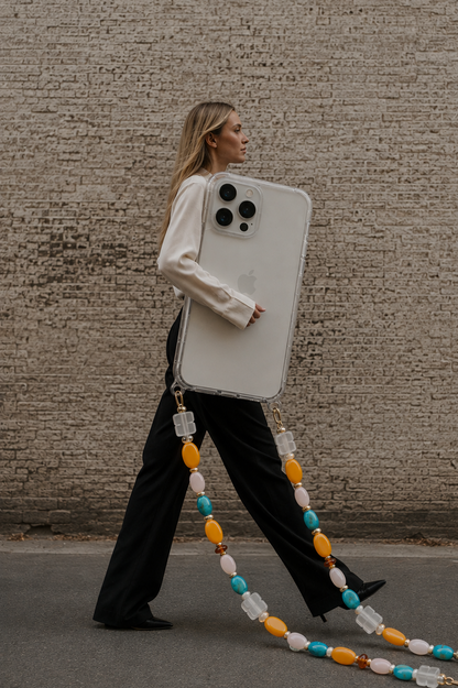 A woman walks past a textured concrete wall carrying a very large white smartphone case with a long decorative beaded strap in yellow, turquoise, white, and orange.