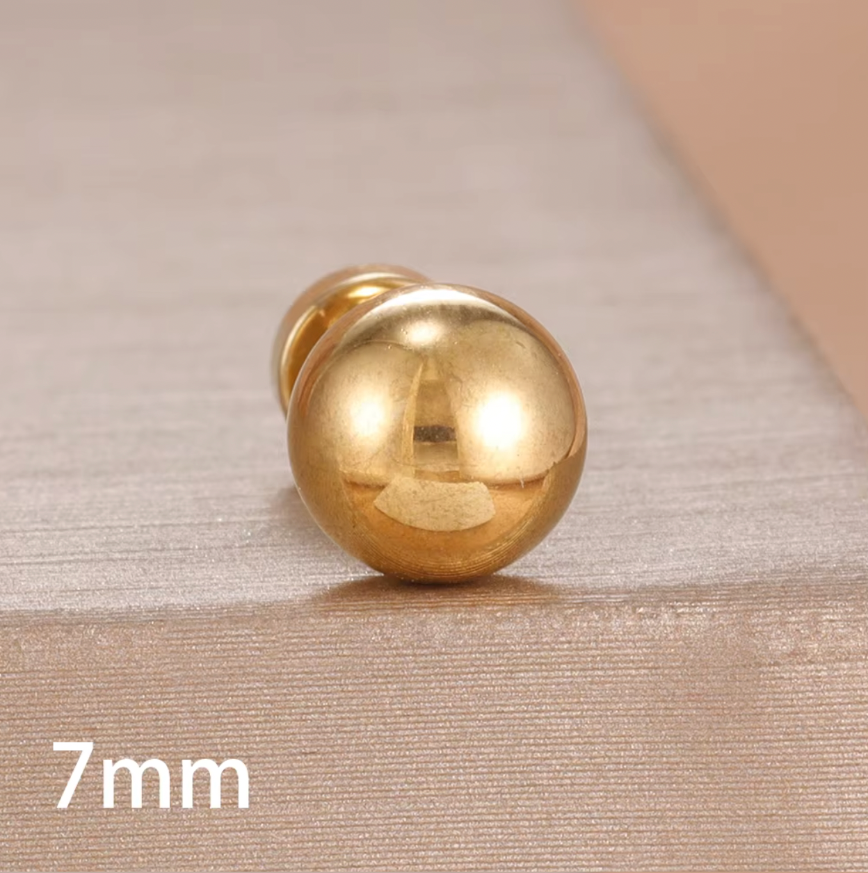 Row of gold ball stud piercings in multiple sizes displayed on a soft fabric background, showing smooth polished finish and flat screw-back closures.
