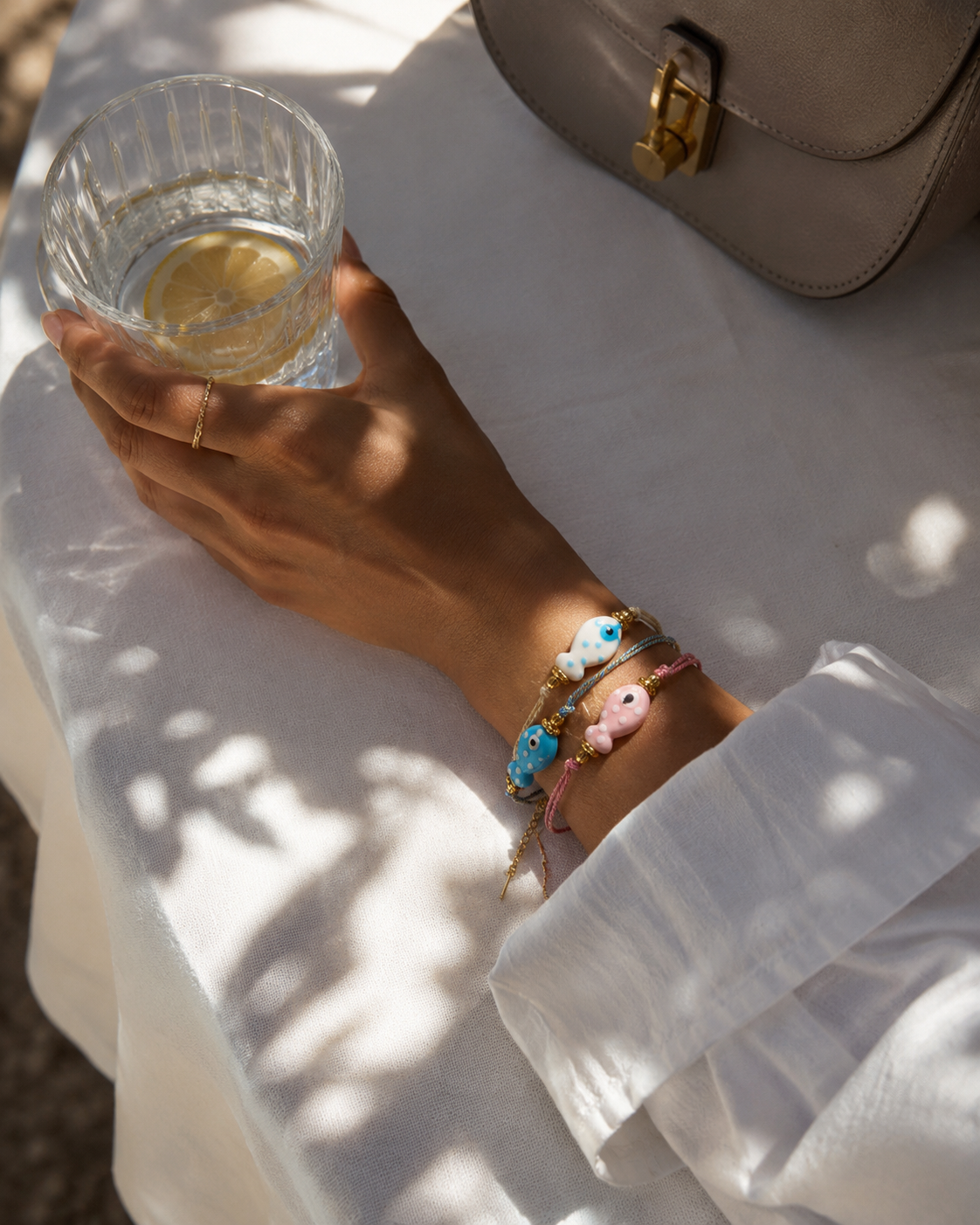 Amalfi BRACELET worn stacked on the wrist with matching fish charms, model holding a glass of water at an outdoor table.