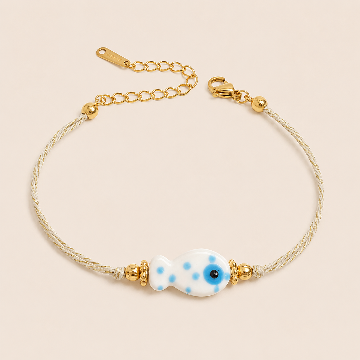 Amalfi BRACELET in white with a ceramic blue polka-dot fish charm on a twisted gold and cream cord, shown flat.