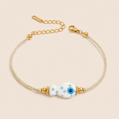 Amalfi BRACELET in white with a ceramic blue polka-dot fish charm on a twisted gold and cream cord, shown flat.