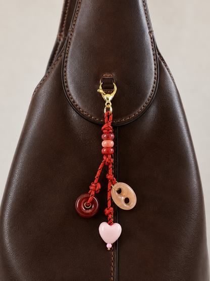 Folie à Trois Amara BAG CHARM, a red beaded cord with heart and agate donut charms, clipped to a dark brown leather bag.