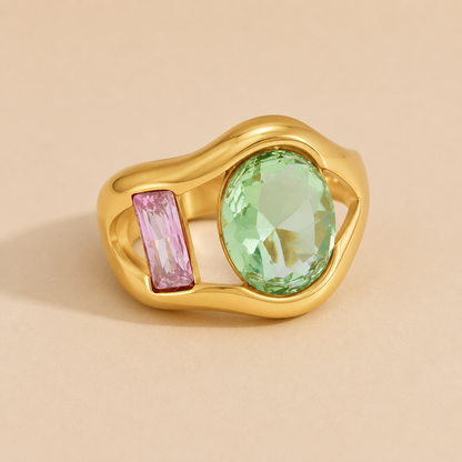 Gold ring set with a green oval stone and a pink baguette-cut stone side by side, on a beige backdrop — Folie à Trois Amara RING.