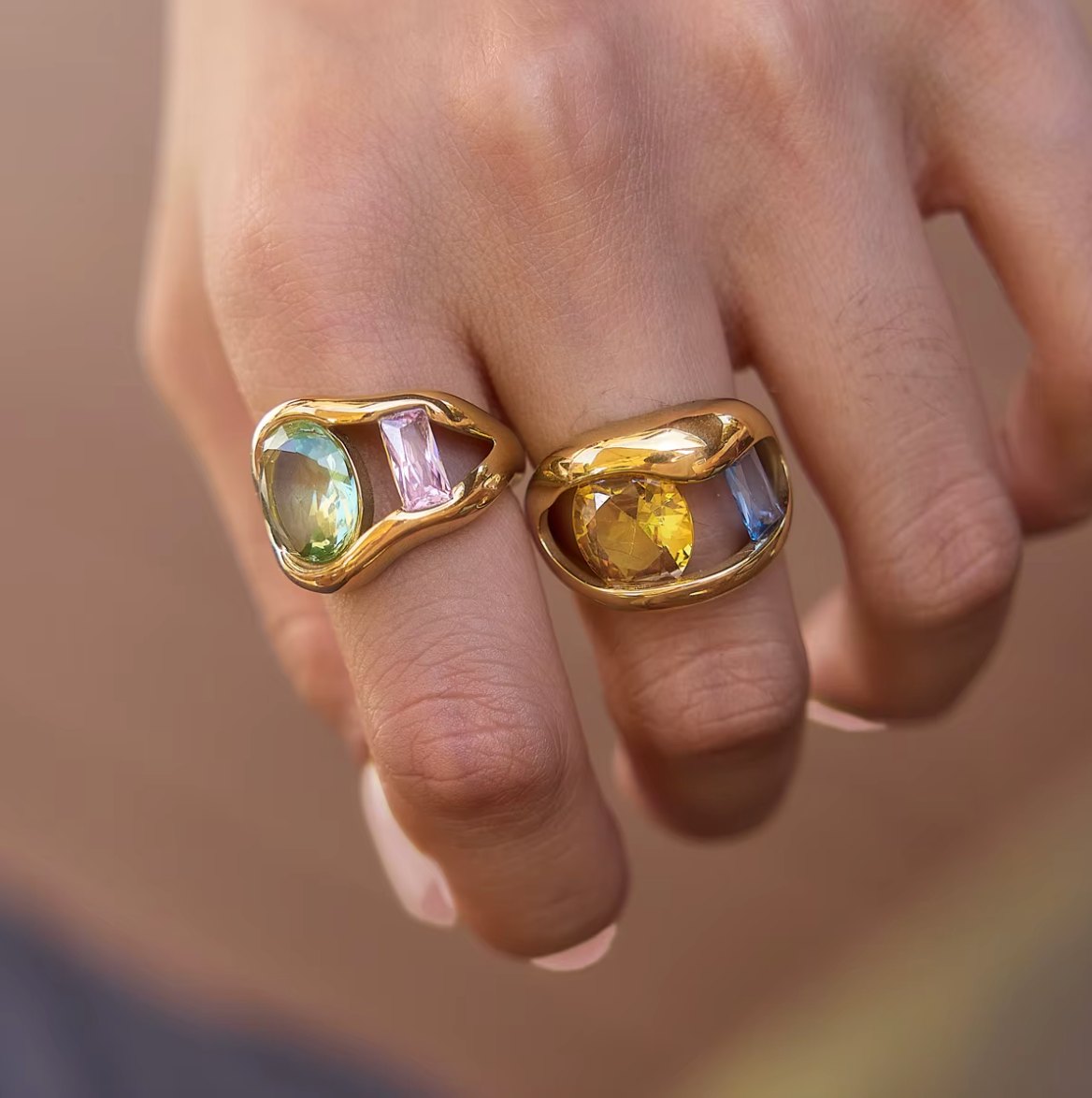 Close-up of two Amara RING colorways stacked on a model's finger, one set with green and pink stones, the other with yellow and blue stones.