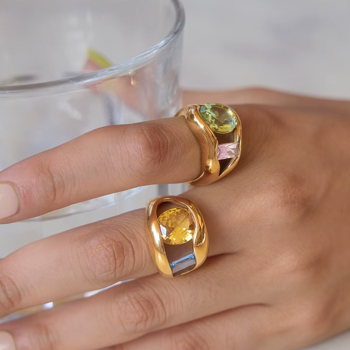Model wears two Amara RING colorways side by side near a glass of water, showing the green-pink and yellow-blue stone pairings up close.