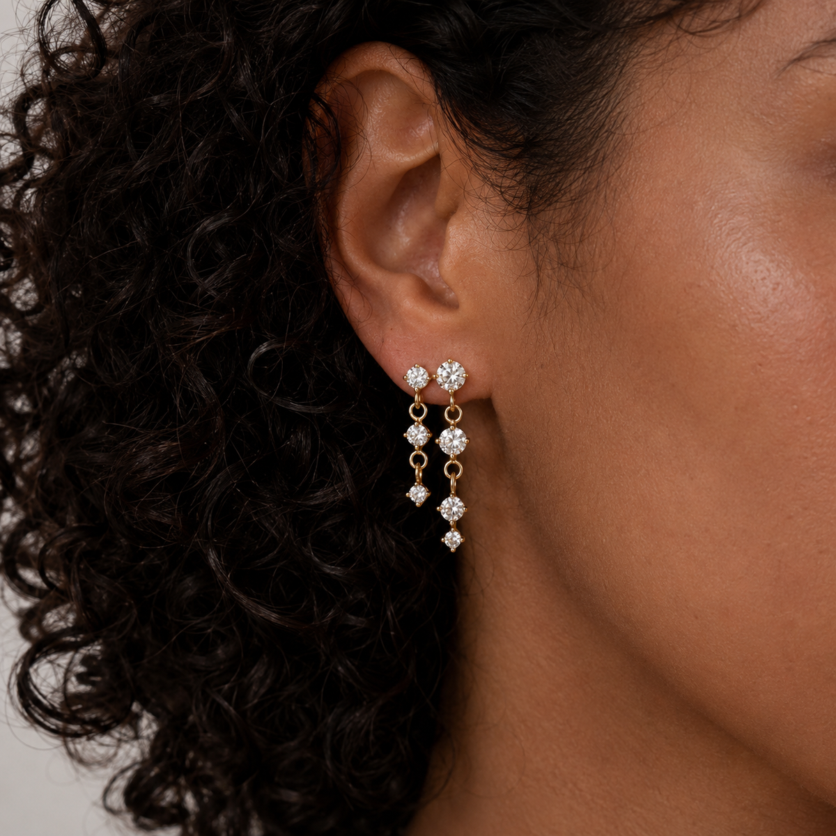 A model wears the gold Andromeda EARRINGS with three graduated round cubic zirconia stones dangling from her ear.