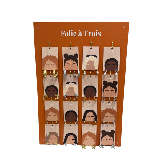 Board With Jewelry display holding 16 pairs of gold and silver earrings clipped to illustrated cards on a terracotta Folie à Trois panel.