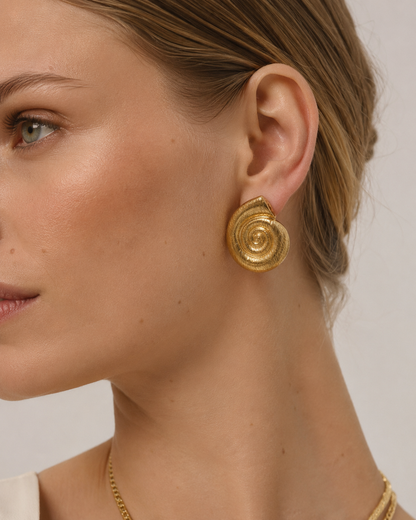 Model wears the Folie à Trois Bonifacio EARRINGS, a brushed gold spiral shell-shaped stud, with a layered gold necklace.