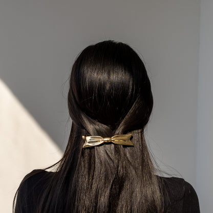 Bow HAIR CLIP in gold-tone metal, worn centered on straight dark hair, back view in sunlight.
