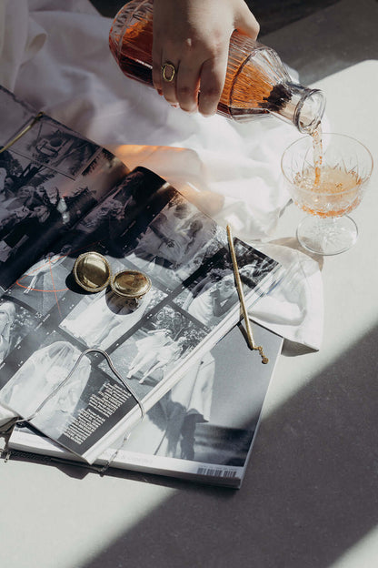 Braided Babe BRACELET styled lifestyle shot of gold cufflinks and a pouring rose drink on a vintage magazine.