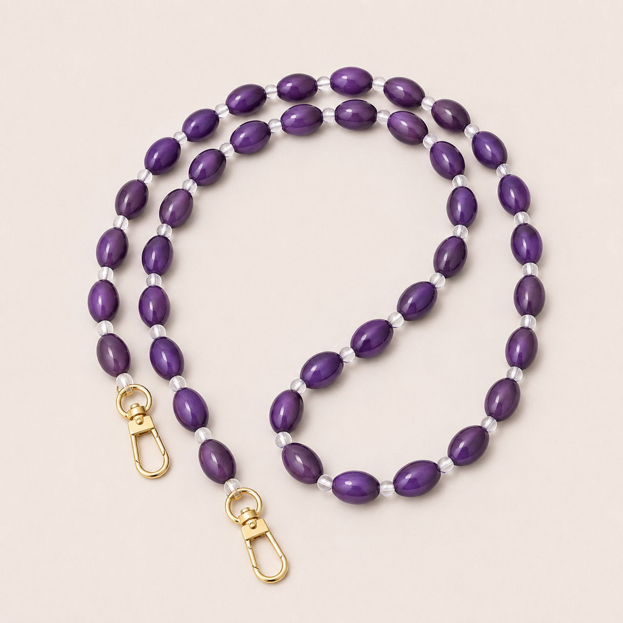 Folie à Trois Bramble long wholesale phone chain in purple oval beads with clear glass spacers and gold clasps, coiled on a cream background.