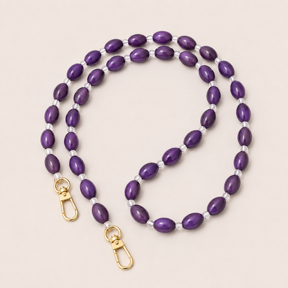 Folie à Trois Bramble long wholesale phone chain in purple oval beads with clear glass spacers and gold clasps, coiled on a cream background.