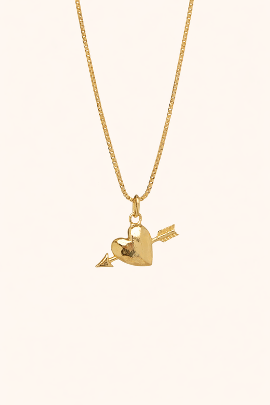 The Burning Hearts NECKLACE in gold, a puffed heart pierced by an arrow charm, hung from a box chain on a cream background.