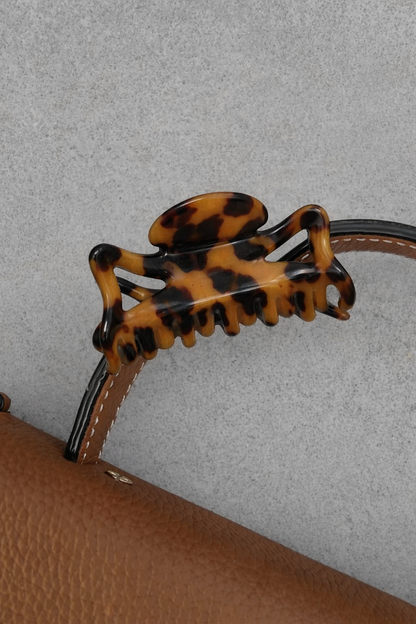 Capri HAIR CLIP's tortoiseshell claw clip styled clipped onto a tan leather bag strap against a grey wall.