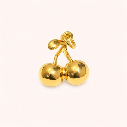 The Cherry CHARM in a studio flat lay, a polished gold pendant shaped like a pair of cherries on a cream background.