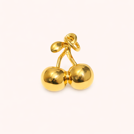 The Cherry CHARM in a studio flat lay, a polished gold pendant shaped like a pair of cherries on a cream background.