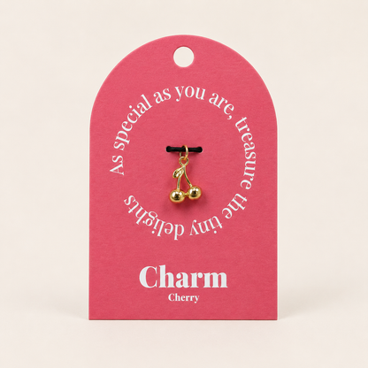 The Cherry CHARM displayed on a coral pink Charm gift tag, the gold cherry pendant hanging from a black cord.