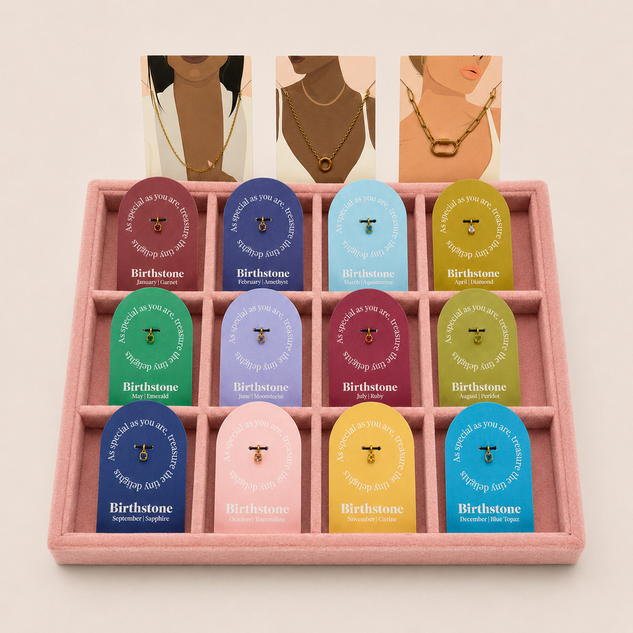 A pink jewelry gift box with 12 individual pouches arranged in a grid, each pouch featuring a different pastel color and the Birthstone brand name. Above the box, three illustrated necklaces on a model's neck are shown as styling suggestions on a warm off-white background.