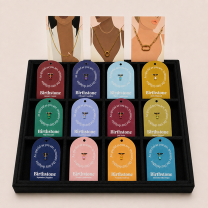 A black jewelry gift box containing 12 colorful jewelry tags, each with a different pastel color and a small gold pendant shown inside. Above the box, three illustrated necklaces on a model's neck are displayed against a light beige background.