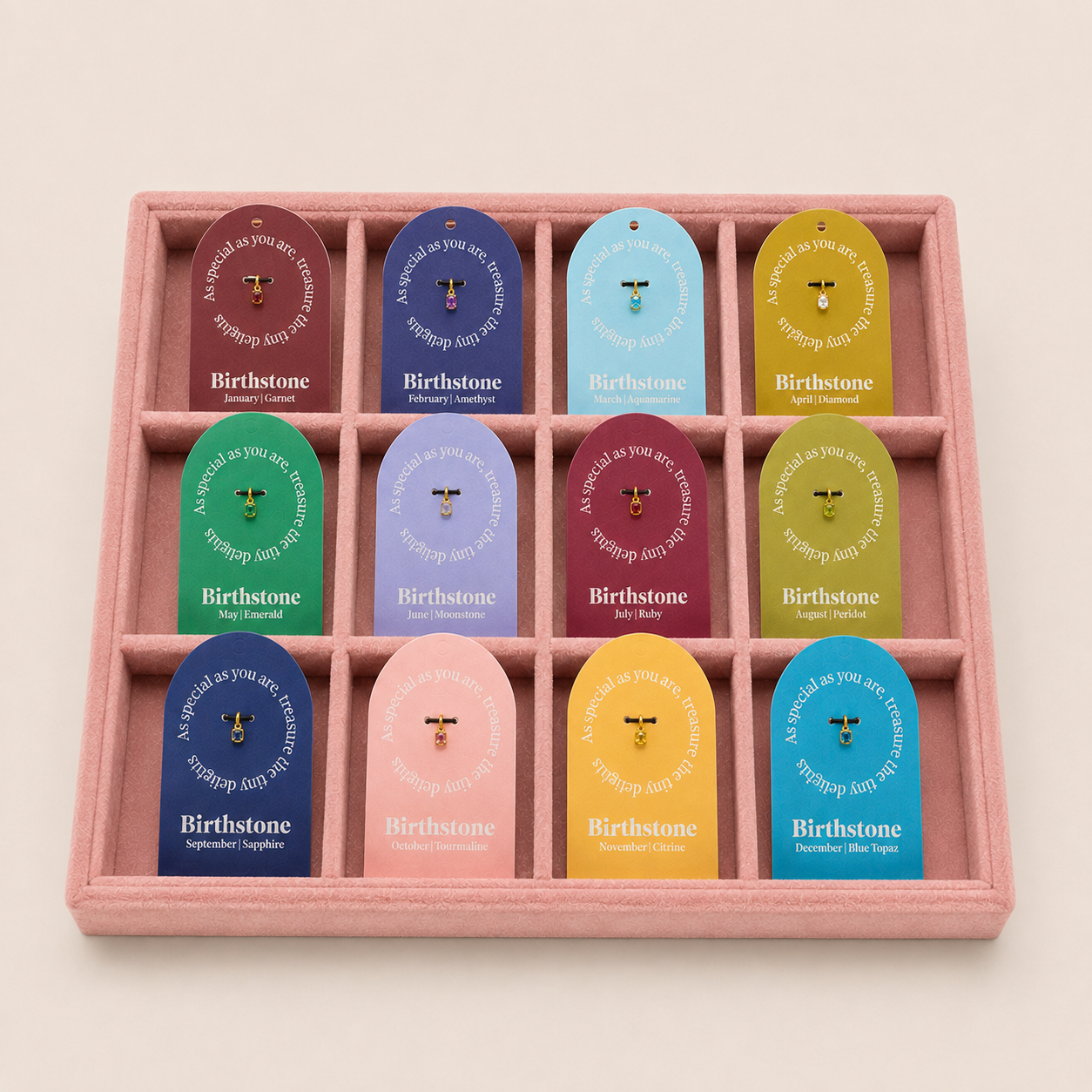 A pink felt jewelry display tray holding twelve small gemstone pendants, each attached to a colorful branded card. The cards read Birthstone and show different gemstone colors: burgundy, blue, light blue, yellow, green, lavender, burgundy, pink, navy, yellow, and light blue.
