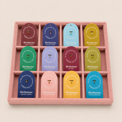 A pink felt jewelry display tray holding twelve small gemstone pendants, each attached to a colorful branded card. The cards read Birthstone and show different gemstone colors: burgundy, blue, light blue, yellow, green, lavender, burgundy, pink, navy, yellow, and light blue.