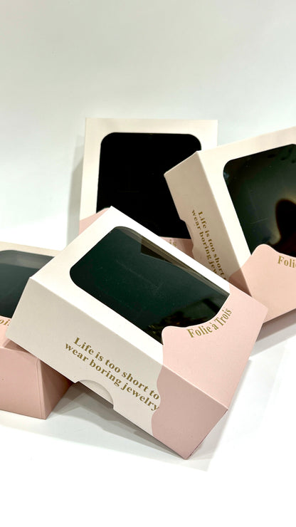 Several pink and cream Folie à Trois clear gift boxes scattered together with the tagline life is too short to wear boring jewelry.