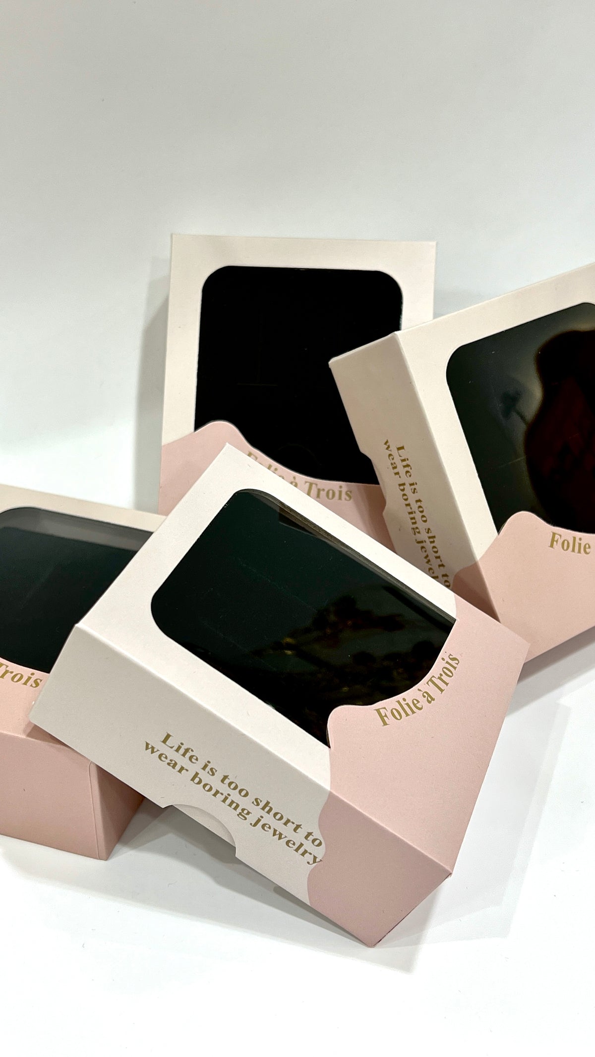 Pink and cream clear-window gift boxes from Folie à Trois stacked at an angle, showing the brand name in gold foil.