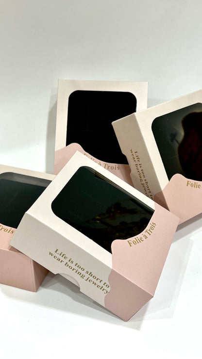 Pink and cream clear-window gift boxes from Folie à Trois stacked at an angle, showing the brand name in gold foil.
