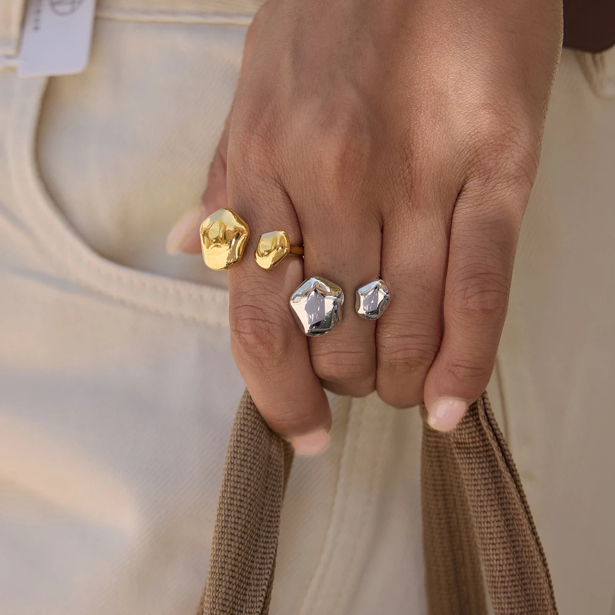 Hand holding a woven bag strap, wearing gold and silver-tone Cloud RINGS against cream trousers — Folie à Trois