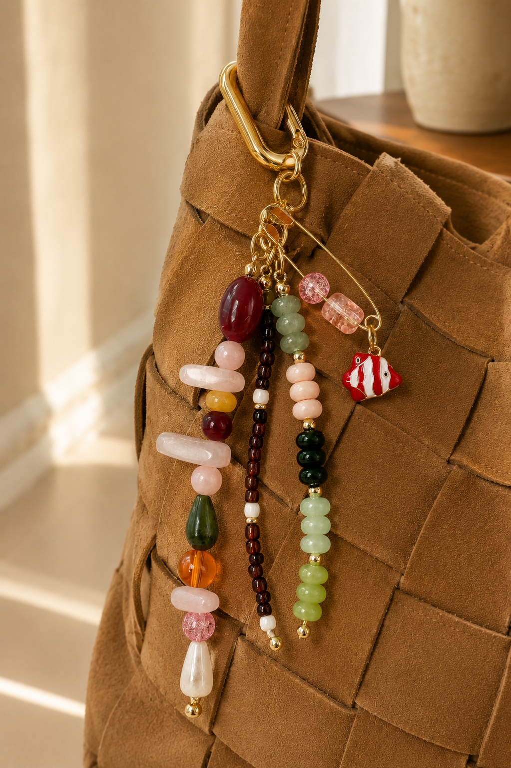Folie à Trois Clownfish BAG CHARM, a red-and-white enamel clownfish charm on a beaded gold pin, clipped to a woven tan tote bag.