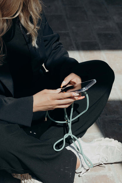 Sage green Colorful PHONE CORD, model seated outdoors holding her phone in sunlight.