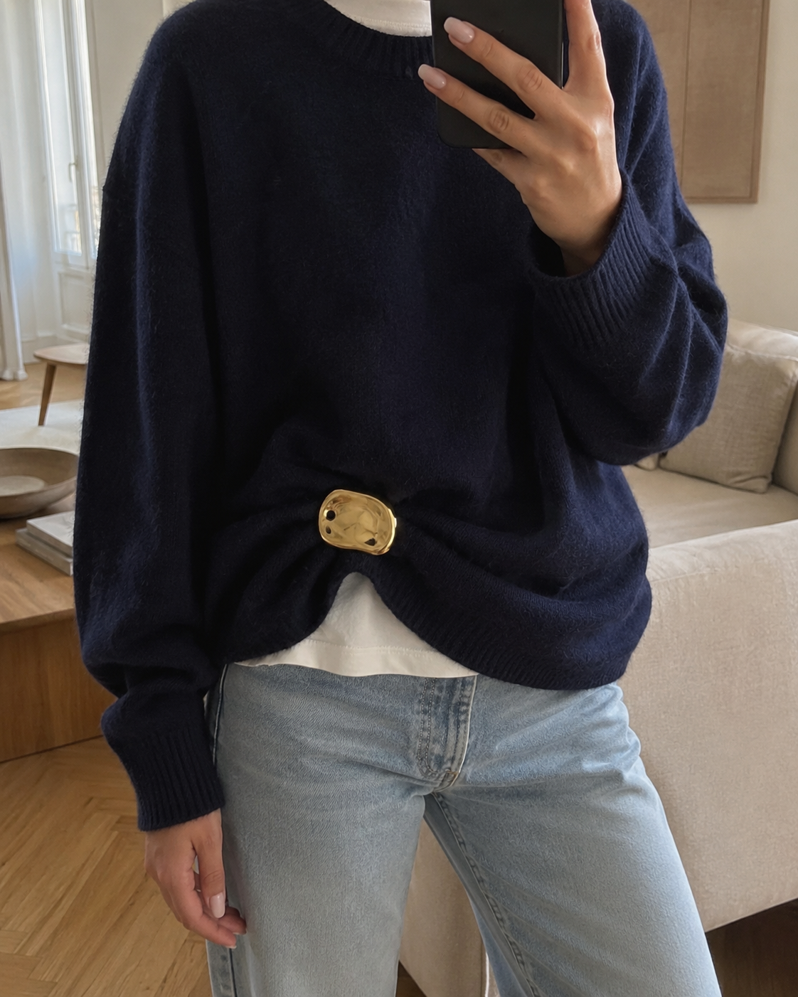 Close-up mirror selfie of a model wearing a gold Costa BROOCH cinching a navy sweater over jeans — Folie à Trois