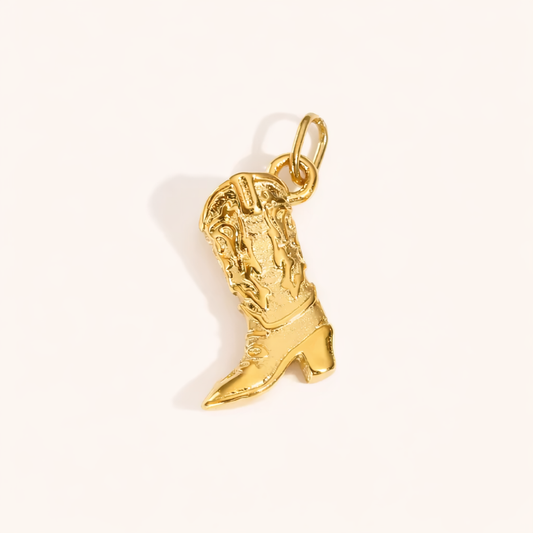 The Cowboy CHARM in a studio flat lay, a textured gold pendant shaped like a western cowboy boot on a cream background.