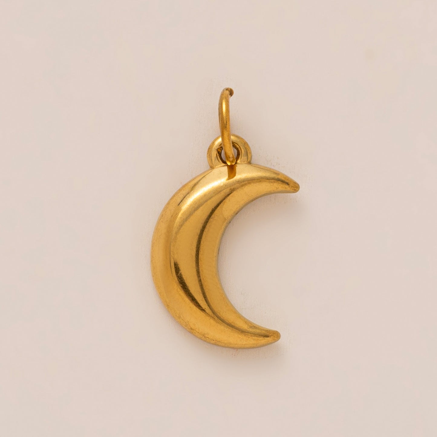 The Crescent Moon CHARM in gold, a smooth curved moon pendant with a jump ring, shown in close-up studio photography on cream.