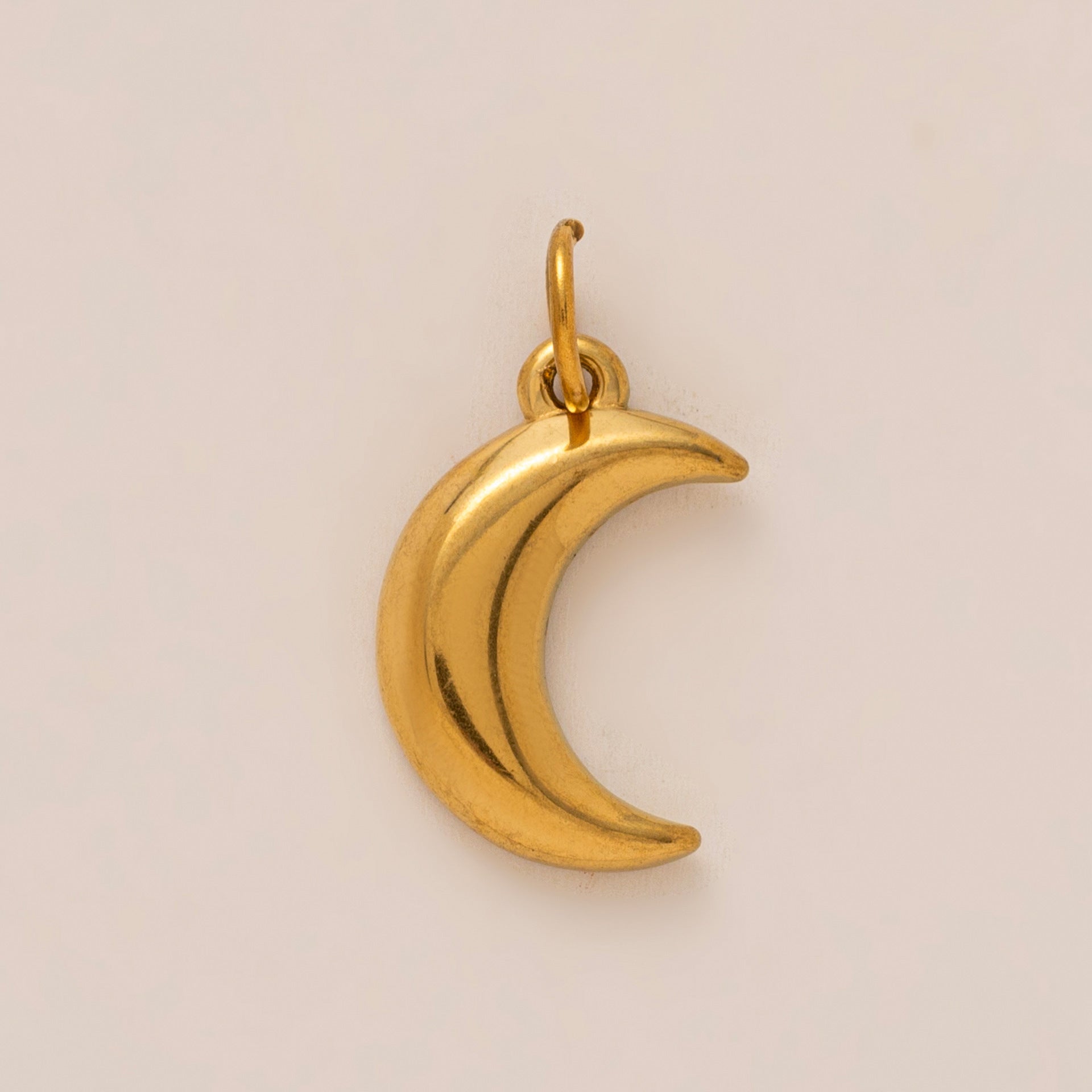 The Crescent Moon CHARM in gold, a smooth curved moon pendant with a jump ring, shown in close-up studio photography on cream.