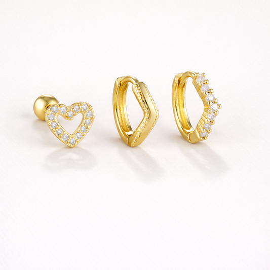 Delia Set of gold jewelry, a pave heart stud, a wavy plain huggie hoop, and a pave chevron hoop, laid flat.