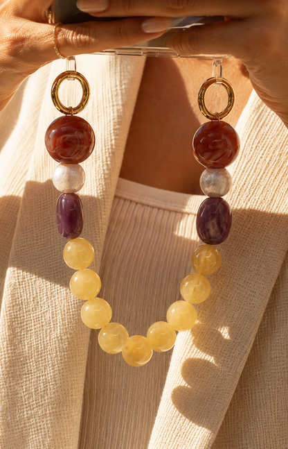 Sunlit close-up of a woman in a cream blazer holding her phone by a red, purple, and yellow bead chain — Folie à Trois Delphine phone chain