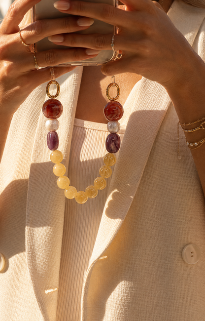 Woman in a cream blazer holds her phone by a red, pearl, purple, and yellow bead chain in golden light — Folie à Trois Delphine phone chain